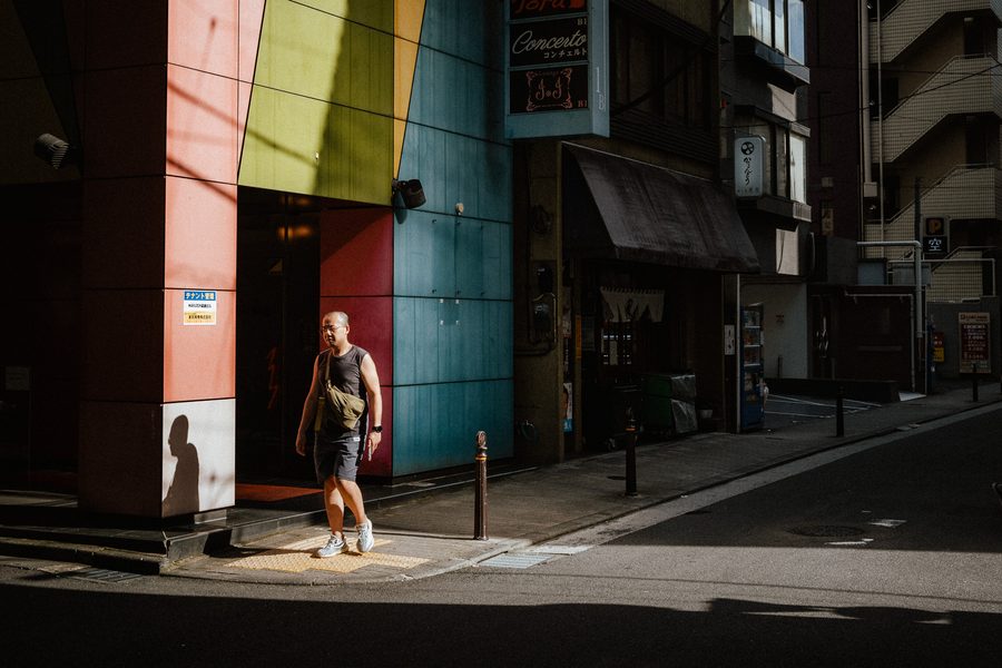 Passante attraversa una lama di luce all’angolo di un edificio con pannelli colorati, ombre nette, Tokyo