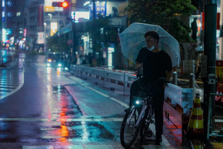 Uomo con ombrello trasparente accanto alla bicicletta su una strada bagnata piena di riflessi, notte, Tokyo