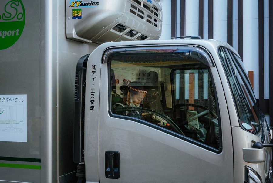 Autista con la fascia annodata in testa al volante di un camion frigo, riflessi delle insegne sul vetro, Shinjuku, Tokyo