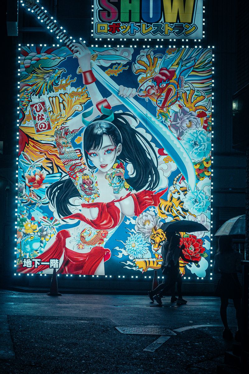 Passanti in silhouette davanti a un grande cartellone illustrato e illuminato, Kabukichō, Tokyo