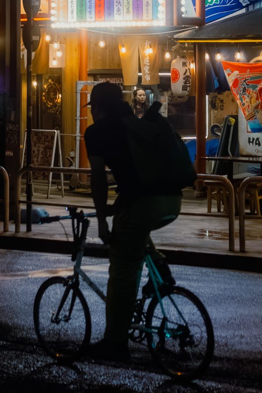Silhouette di un ciclista davanti a un mercato izakaya illuminato, notte di pioggia, Shimbashi, Tokyo