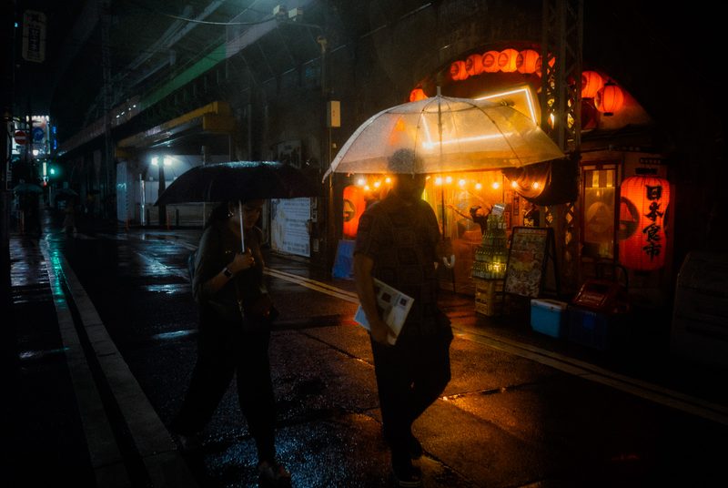 Coppia con ombrelli attraversa davanti alle lanterne rosse di un mercato izakaya, notte di pioggia, Shimbashi, Tokyo