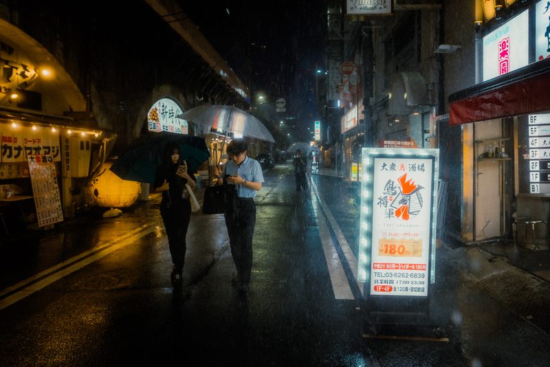 Coppia con ombrelli davanti all'insegna luminosa dell'izakaya Tori Shougun sotto la pioggia battente, Shimbashi, Tokyo