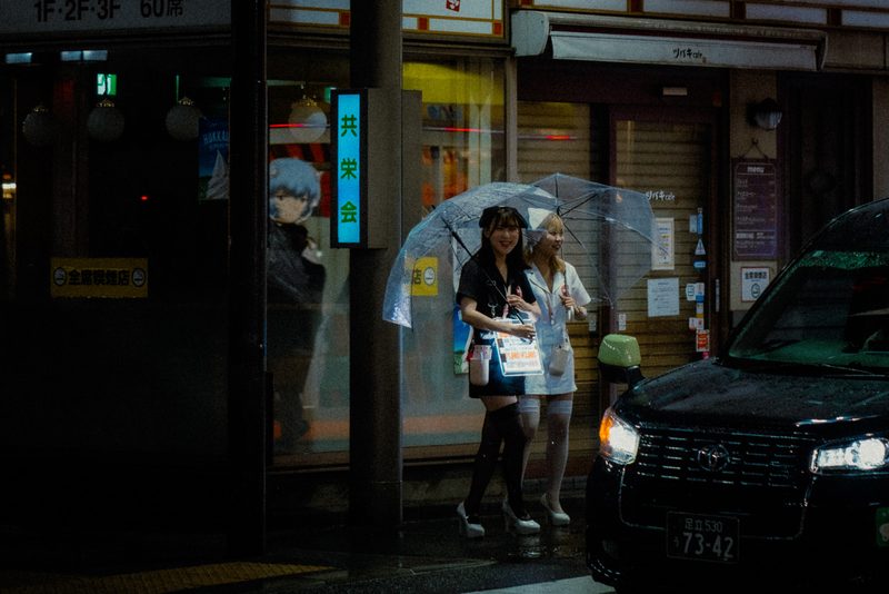Due ragazze in costume con ombrelli trasparenti accanto a un'auto nera, notte di pioggia, Shimbashi, Tokyo