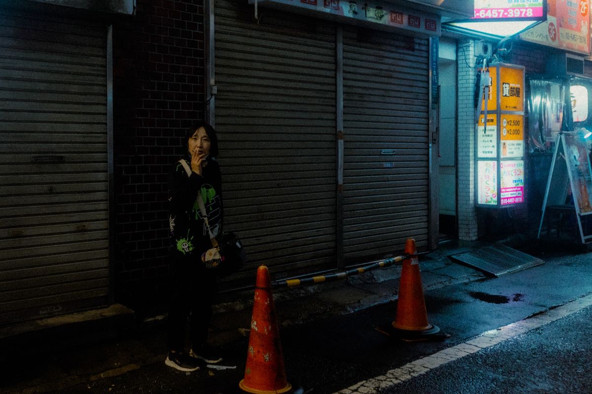 Donna fuma appoggiata alle serrande chiuse tra due coni stradali, Kabukichō, Tokyo