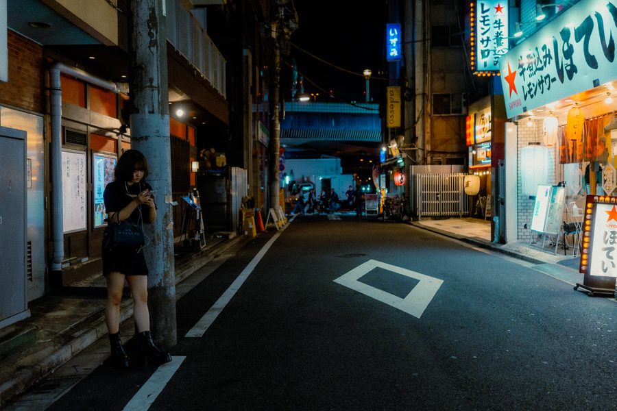 Donna appoggiata a un palo guarda il telefono in una strada deserta di notte, insegne di izakaya sulla destra, Tokyo