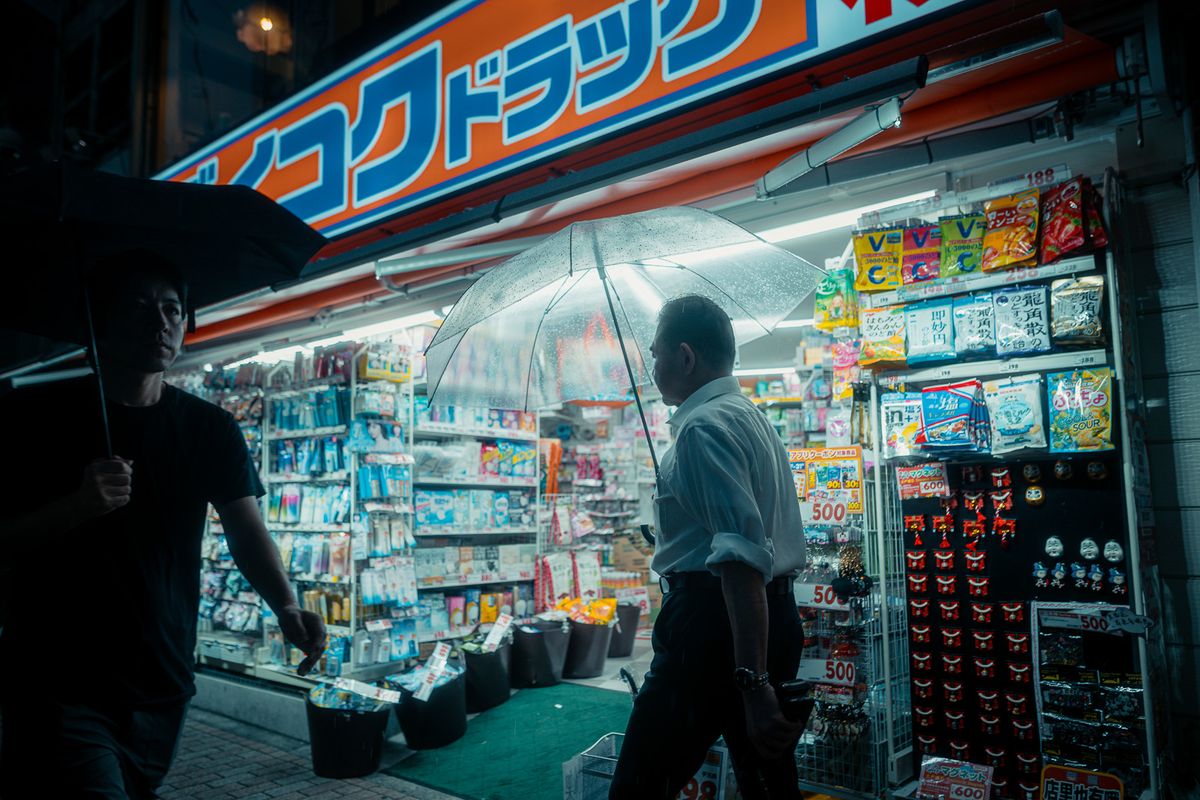 Due uomini con l’ombrello si incrociano davanti a una farmacia illuminata, Shinjuku, Tokyo