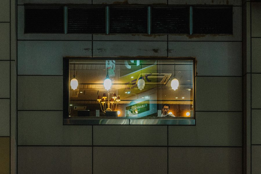Unica finestra illuminata in una facciata buia, con lampadari accesi e una figura all’interno, Tokyo
