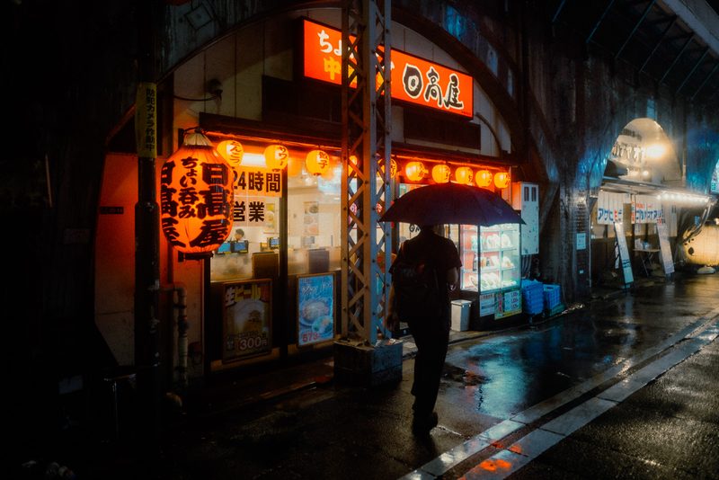 Uomo con ombrello davanti al Hidakaya aperto 24 ore con grande lanterna rossa sotto un arco ferroviario, notte di pioggia, Tokyo