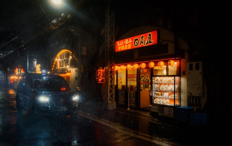 Taxi coi fari accesi sotto un arco ferroviario accanto al Hidakaya illuminato, notte di pioggia, Tokyo