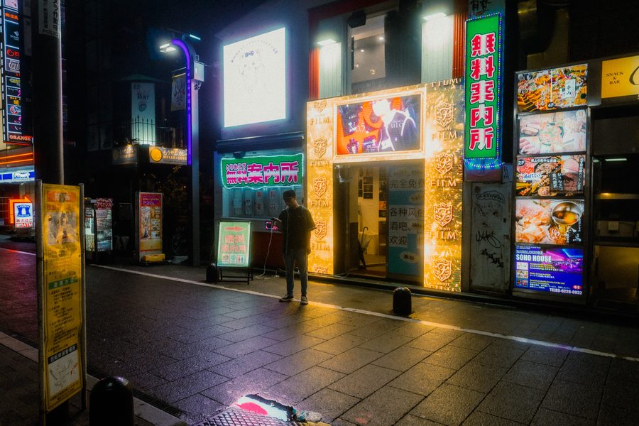 Un uomo solo al telefono davanti alle insegne di un ufficio informazioni, marciapiede bagnato, Kabukichō, Tokyo