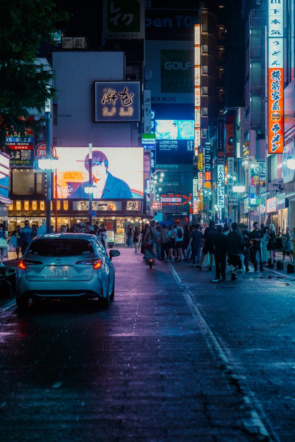 Auto bianca ferma sull’asfalto bagnato mentre la folla scorre sul marciapiede tra le insegne, Kabukichō di notte, Tokyo