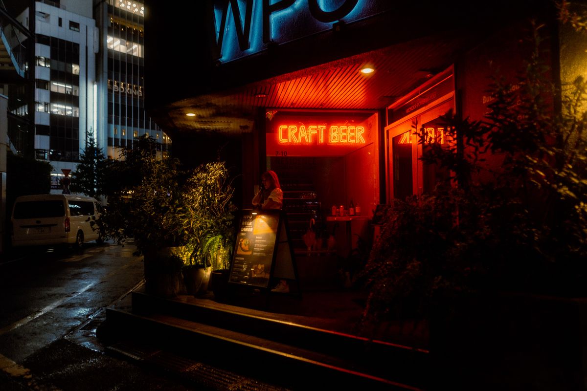 Ragazza sulla soglia di un locale sotto l’insegna al neon rossa Craft Beer, Shinjuku, Tokyo