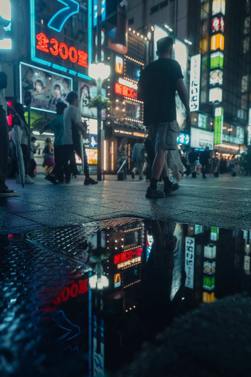 Passante e insegne riflessi in una pozzanghera dopo la pioggia, Kabukichō, Tokyo