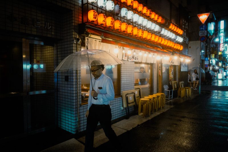 Uomo in camicia con ombrello trasparente davanti a un muro di lanterne rosse e bianche, notte di pioggia, Tokyo