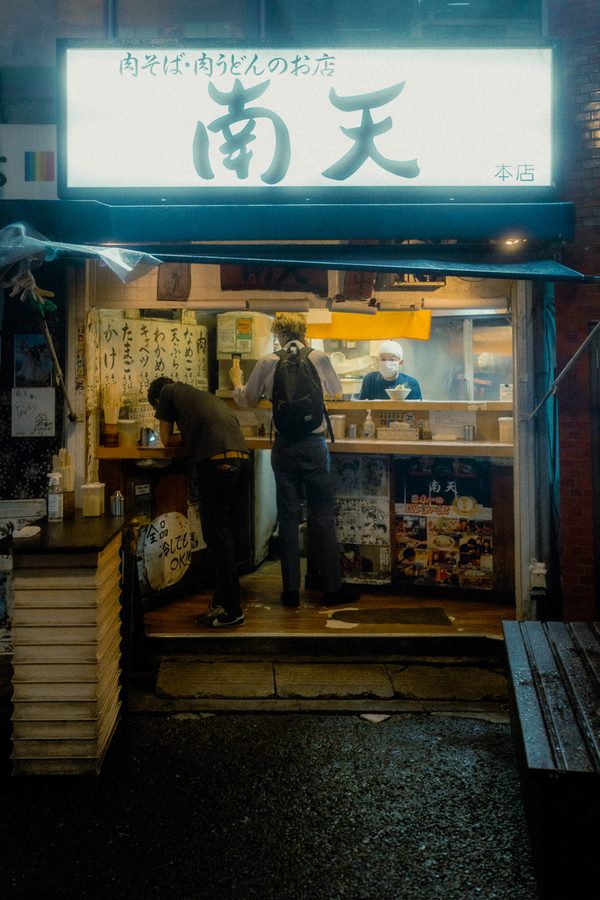 Due clienti al banco di una tavola calda di soba sotto l’insegna illuminata Nanten, il cuoco oltre il bancone, di notte a Tokyo