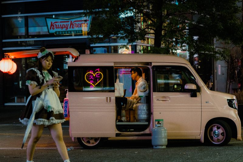 Ragazza in costume da maid davanti a un furgone rosa di baby castella con cuore al neon, di notte a Tokyo