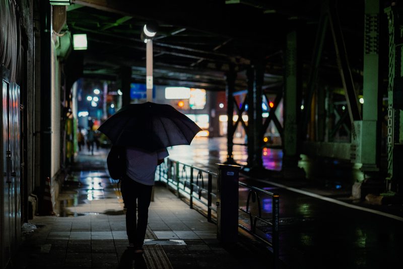 Uomo con ombrello scuro cammina sotto la ferrovia tra luci sfocate, notte di pioggia, Tokyo