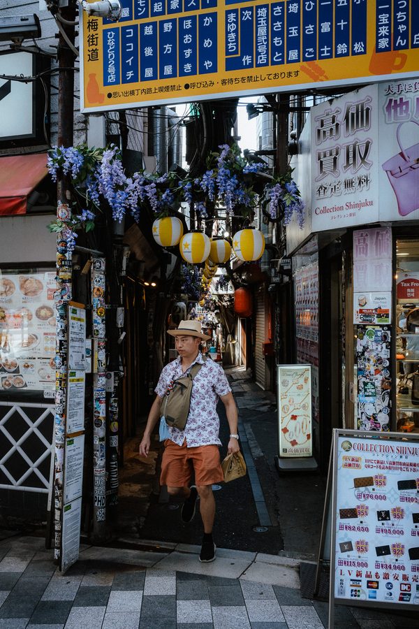 Uomo con cappello di paglia e camicia a fiori esce dal vicolo di Omoide Yokochō sotto lanterne e glicine, Tokyo