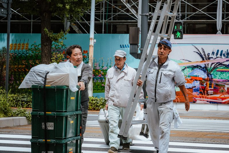 Tre operai attraversano le strisce con una scala e casse davanti a un murale, di giorno, Tokyo