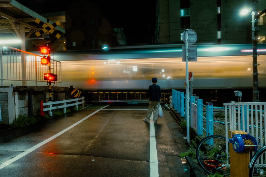 Uomo con una busta della spesa attende al passaggio a livello mentre il treno sfila mosso, notte, Tokyo