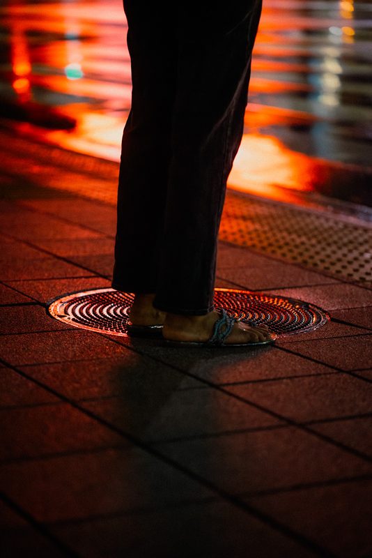 Piedi in sandali su un tombino illuminato con riflessi rossi sull'asfalto bagnato, dettaglio, notte, Tokyo