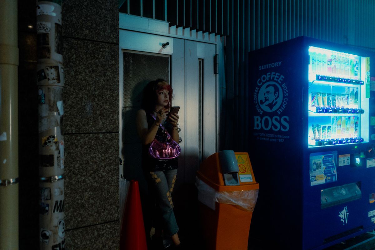 Ragazza col telefono appoggiata a una porta accanto a un distributore automatico illuminato, Tokyo
