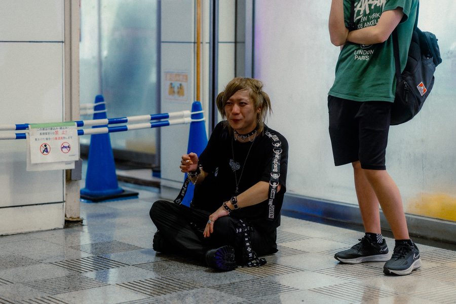 Ragazzo seduto a gambe incrociate a terra accanto al cartello che vieta di sedersi, passante in piedi di fianco, Tokyo