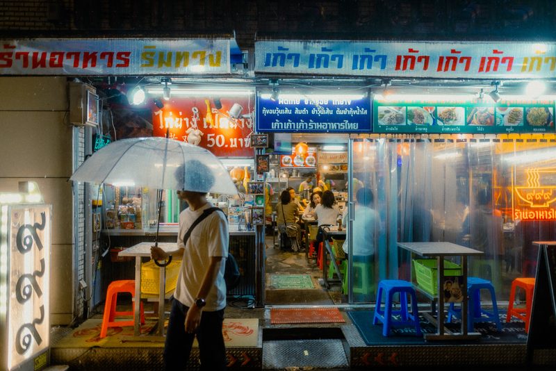 Uomo con ombrello trasparente davanti a un ristorante thailandese illuminato, notte di pioggia, Tokyo