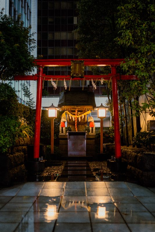 Piccolo santuario Inari con torii rosso e statue di volpe incastonato tra i grattacieli, pavimento bagnato, di notte, Tokyo
