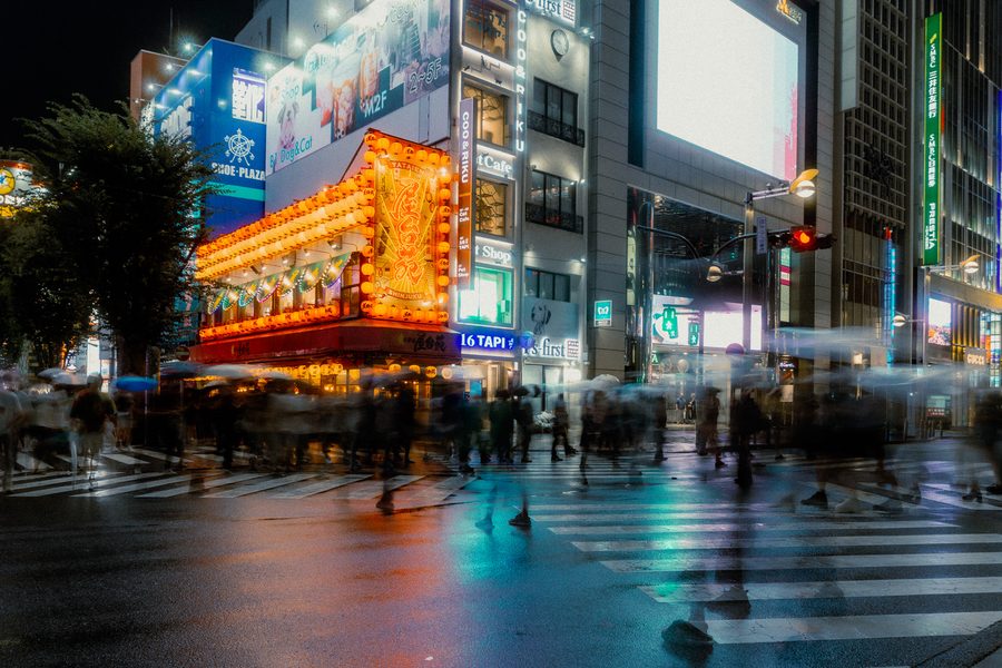 Folla mossa in lunga esposizione attraversa l’incrocio bagnato davanti alle lanterne arancioni di un locale, Shinjuku, Tokyo