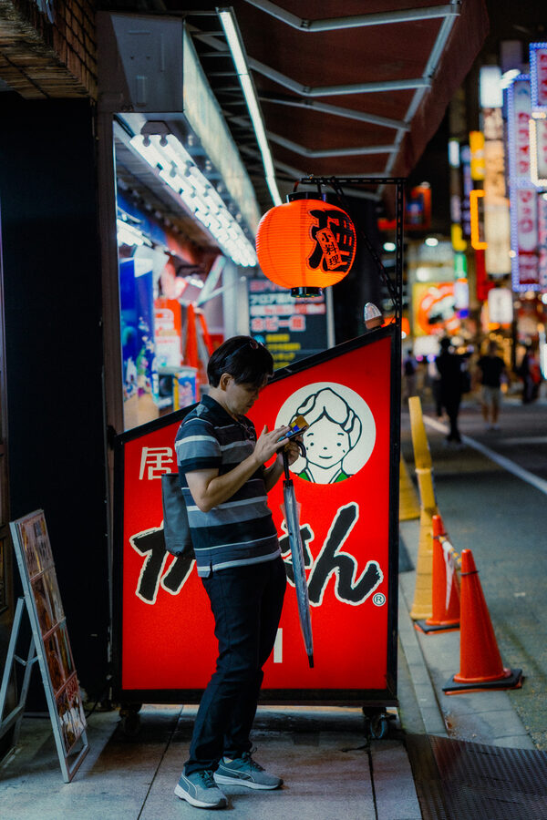 Uomo assorto nel telefono sotto una lanterna rossa, lungo una strada notturna di Shinjuku, Tokyo
