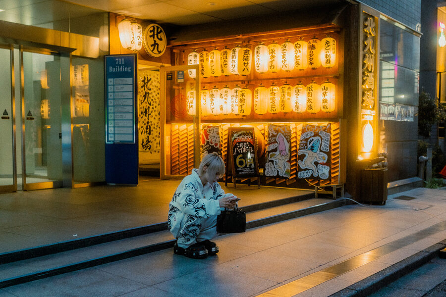 Ragazzo accovacciato guarda il telefono davanti a un locale illuminato da file di lanterne, Shinjuku, Tokyo