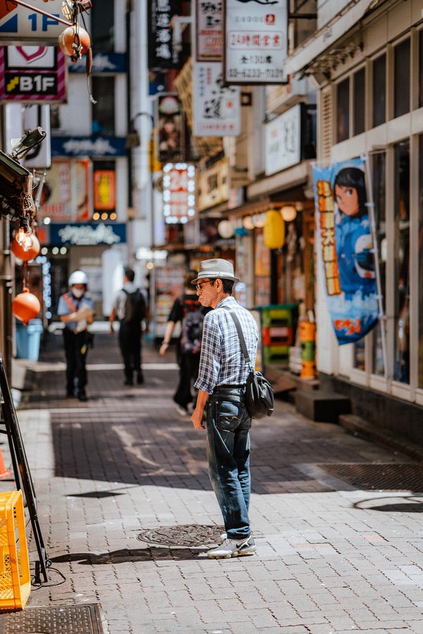 Uomo anziano con cappello di paglia e camicia a quadri fermo in una via commerciale di giorno, Shinjuku, Tokyo