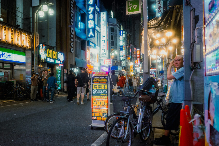 Biciclette e passanti lungo una via fitta di insegne notturne, Shinjuku, Tokyo