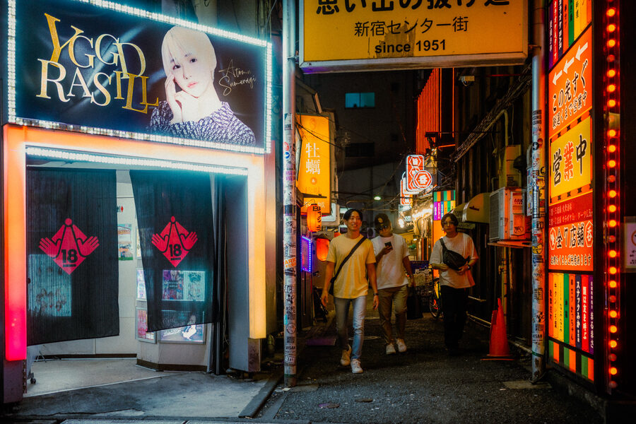 Tre passanti escono da un vicolo stretto tra insegne rosse e bianche, Shinjuku, Tokyo