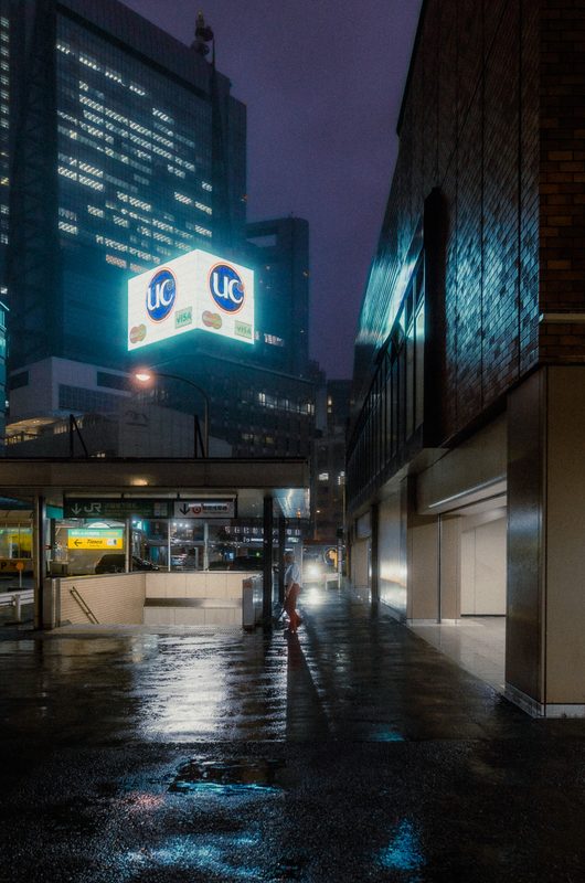 Figura solitaria sotto l'insegna al neon UC nella piazza di Shiodome, notte di pioggia, Tokyo