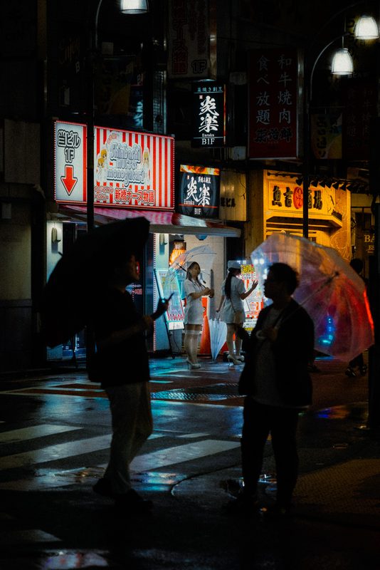 Silhouette di passanti con ombrelli e ragazze in costume da infermiera sullo sfondo, insegne al neon, notte di pioggia, Shimbashi, Tokyo