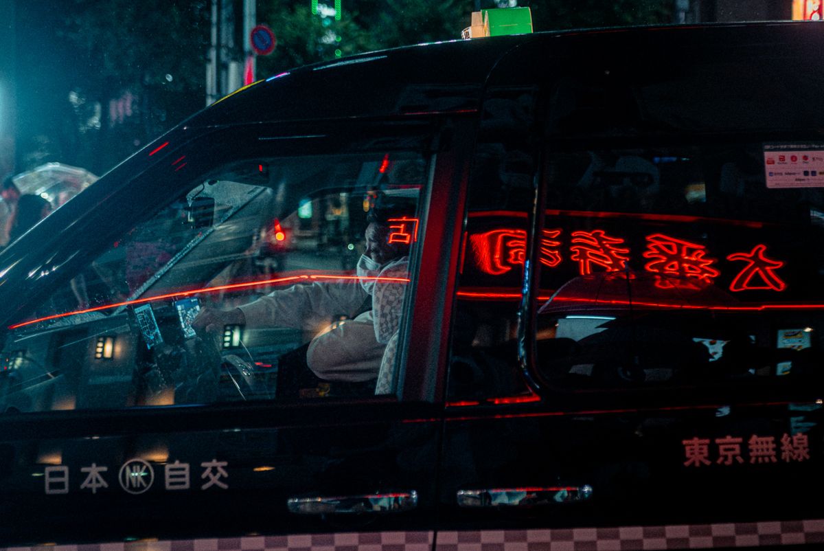 Grande scritta al neon rossa riflessa sulla fiancata di un taxi con l’autista al volante, Tokyo