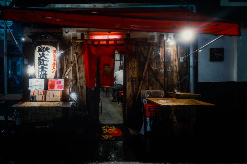 Ingresso in legno dell'izakaya Tekka Bushido con lanterna e tendaggio rosso, un avventore solo all'interno, notte di pioggia, Tokyo