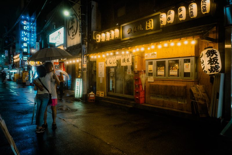 Coppia sotto l'ombrello davanti a un teppanyaki con lanterne e lucine, notte di pioggia, Shimbashi, Tokyo