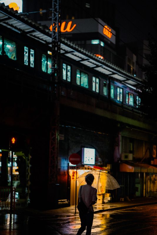 Figura con ombrello trasparente attraversa mentre un treno illuminato passa sul viadotto, notte di pioggia, Tokyo
