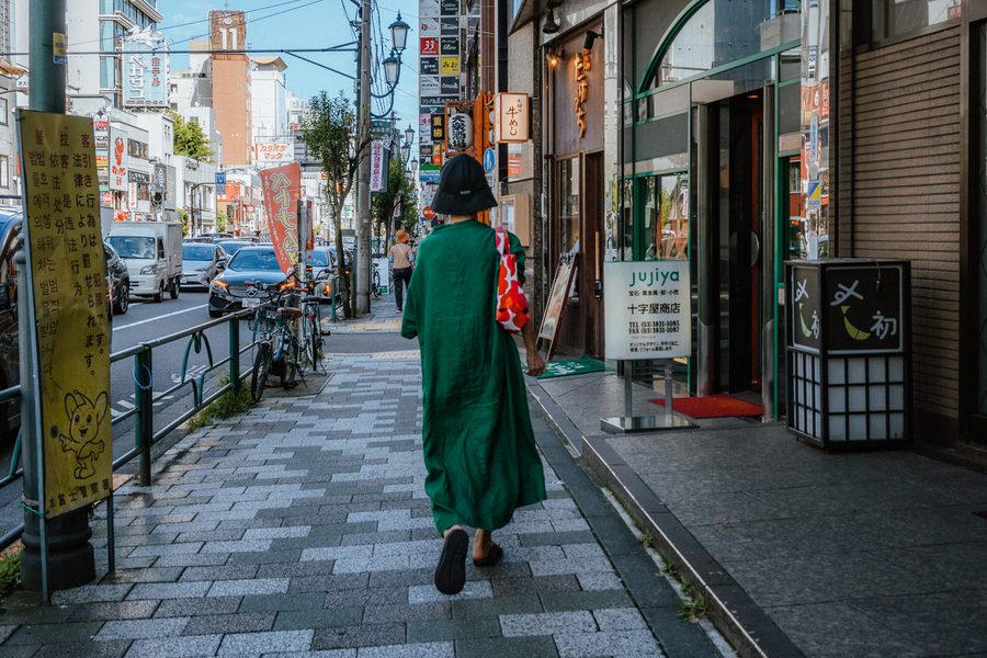 Donna con abito lungo verde e cappello a secchiello cammina di spalle lungo il marciapiede, Ueno, Tokyo