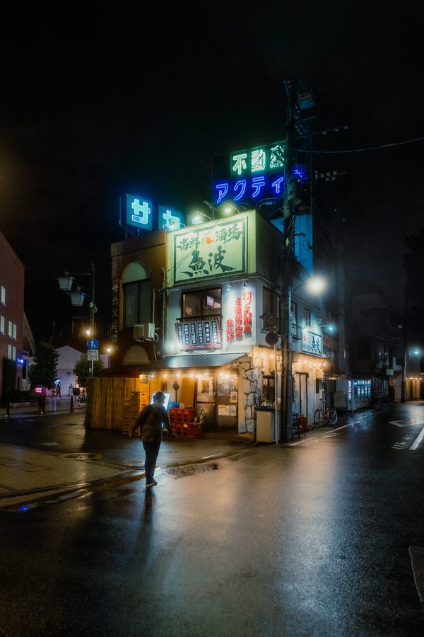 Passante solitario attraversa l’incrocio bagnato davanti all’izakaya Uonami e alle insegne al neon, notte, Tokyo