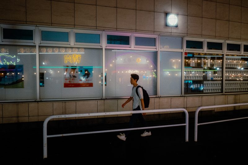 Uomo con zaino cammina davanti a una vetrina al neon di una stazione, di notte, Tokyo
