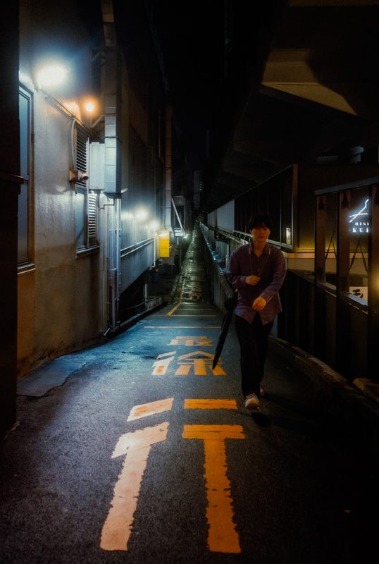 Uomo con ombrello in un lungo vicolo sotto i binari con frecce arancioni sull'asfalto, notte di pioggia, Tokyo