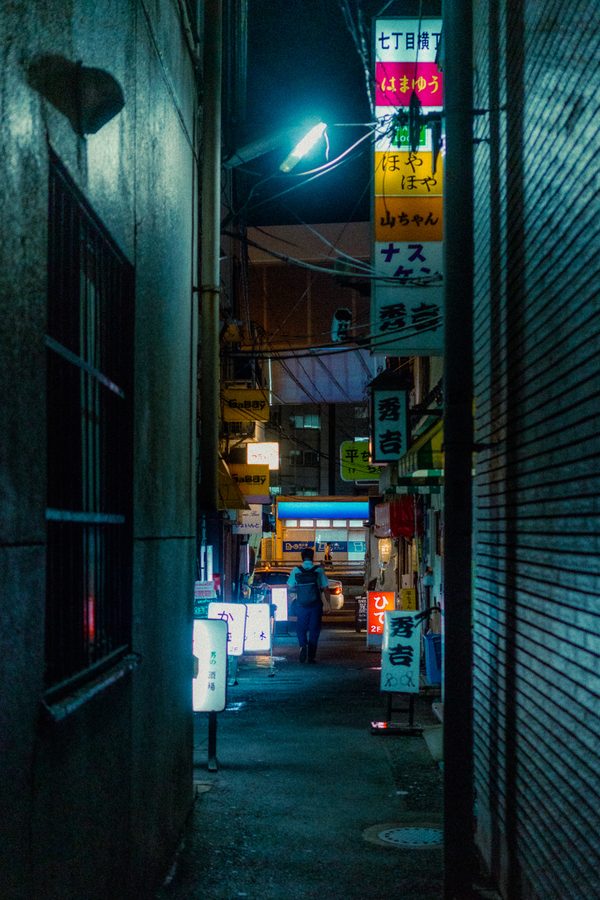 Vicolo stretto di notte incorniciato da due muri, insegne verticali degli izakaya e una figura in fondo, Tokyo