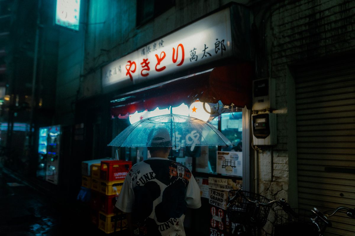 Uomo di spalle con ombrello trasparente davanti a un banco di yakitori, Tokyo