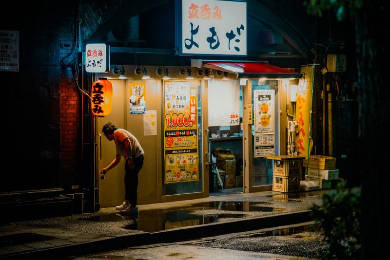 Uomo davanti al bar in piedi Yomoda con lanterna rossa sotto un arco ferroviario, notte di pioggia, Tokyo